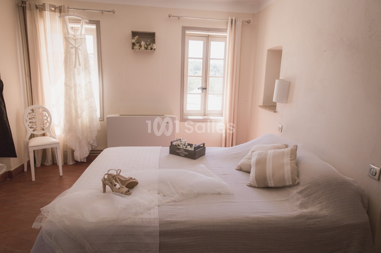 Robe de mariée suspendue près d'une fenêtre dans une chambre lumineuse avec lit décoré d'accessoires de mariage.