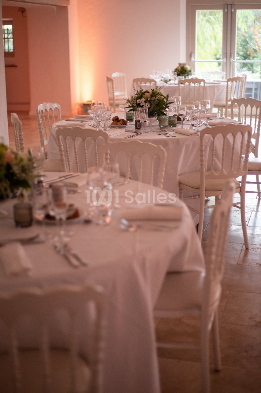 Salle de réception avec des tables rondes dressées, nappes blanches, chaises élégantes et décorations florales.