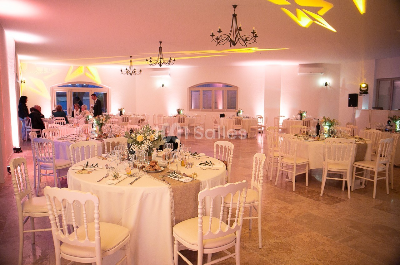 Salle de réception élégamment décorée avec des tables rondes, nappes blanches, chaises assorties et éclairage chaleureux.