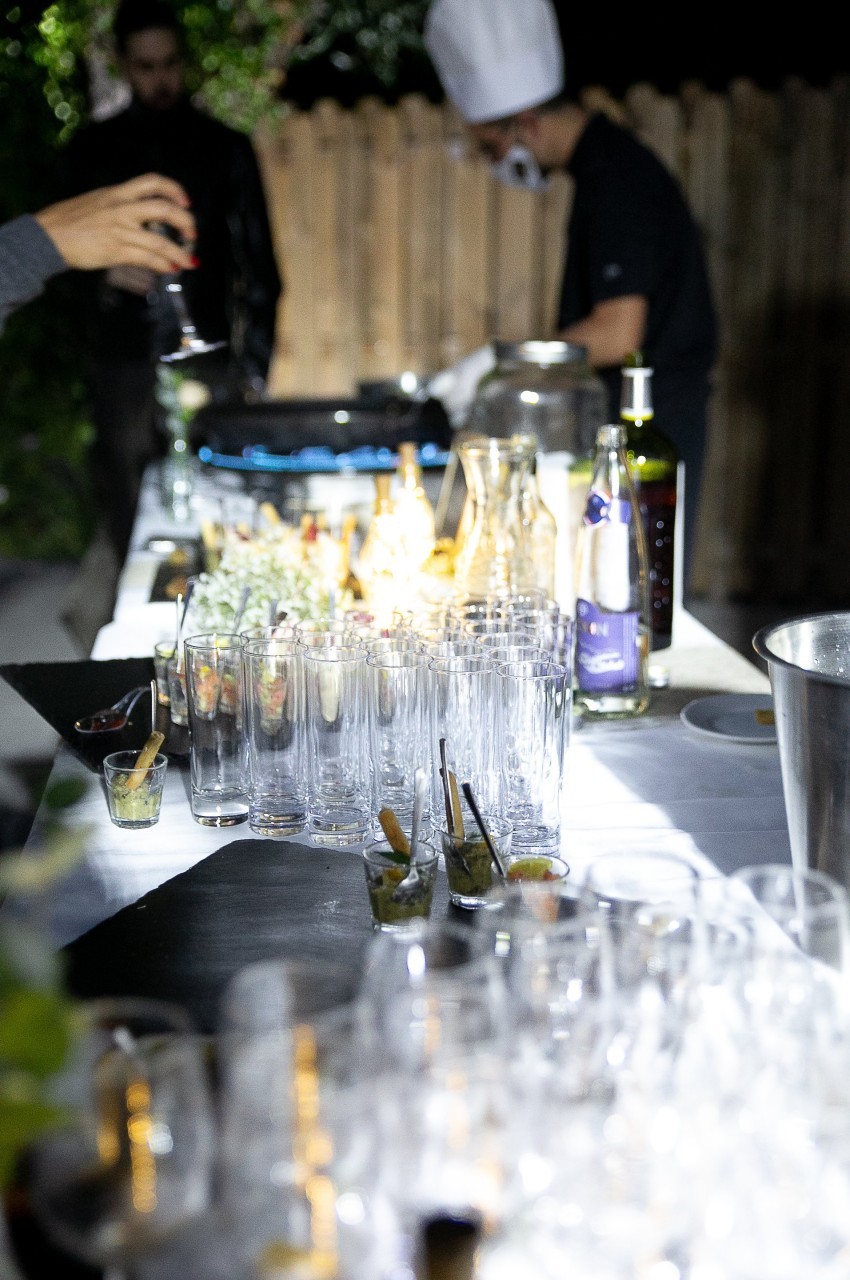 Table de service en extérieur avec verres vides alignés, amuse-bouches et un chef cuisinant en arrière-plan.