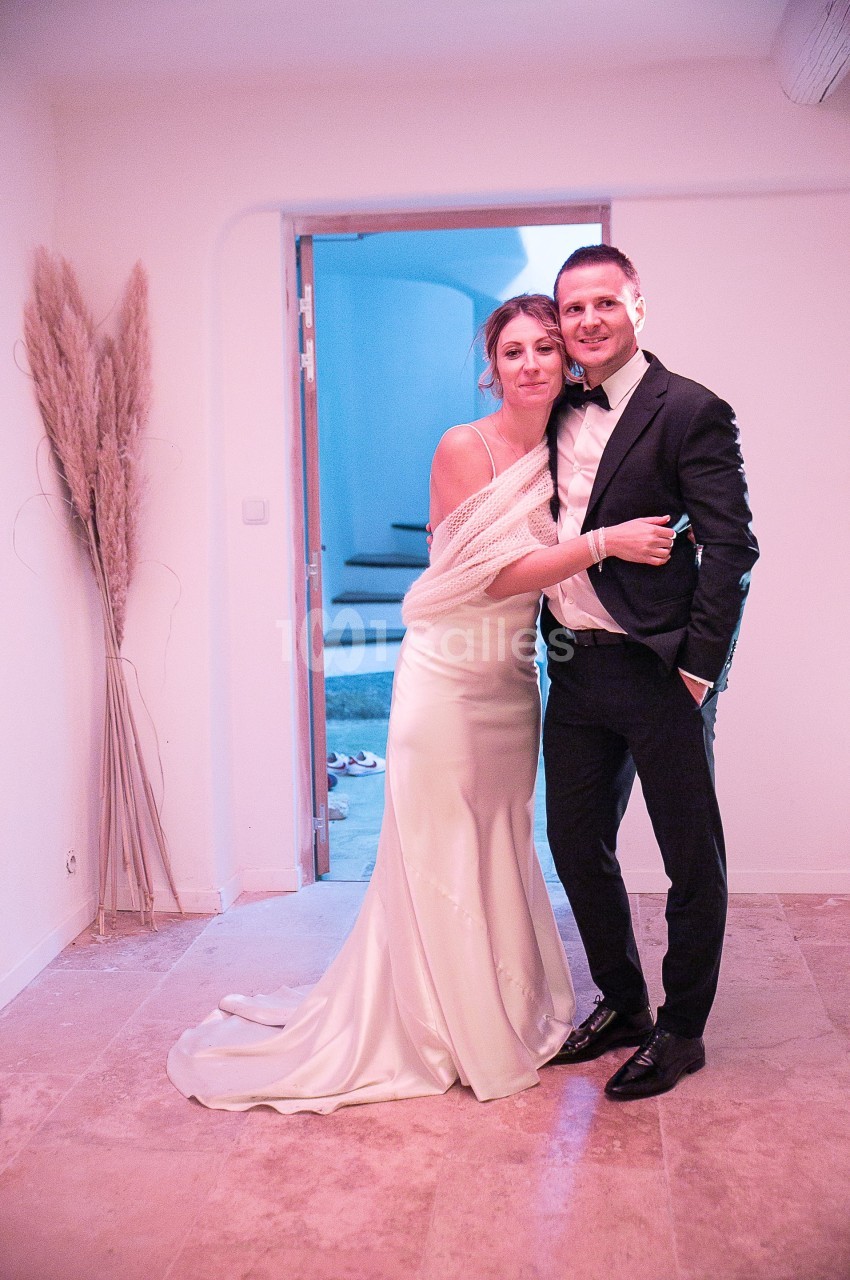 Un couple habillé élégamment, la femme en robe blanche et l'homme en costume, pose dans une pièce lumineuse.