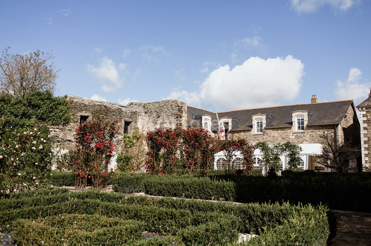 Façade en pierre d'une bâtisse ancienne entourée de rosiers grimpants et d'un jardin structuré.