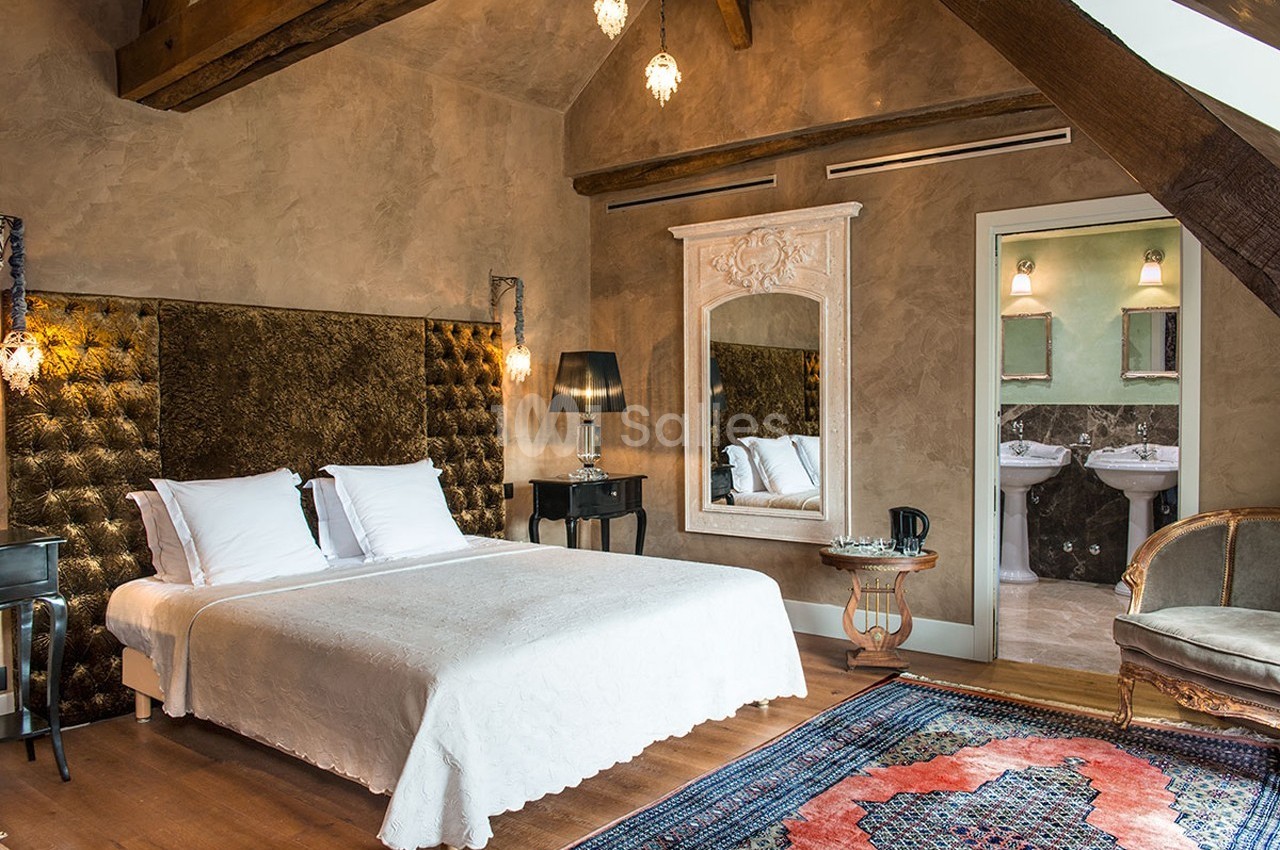 Chambre lumineuse avec lit double, murs texturés, miroir décoratif, poutres apparentes et vue sur une salle de bain…