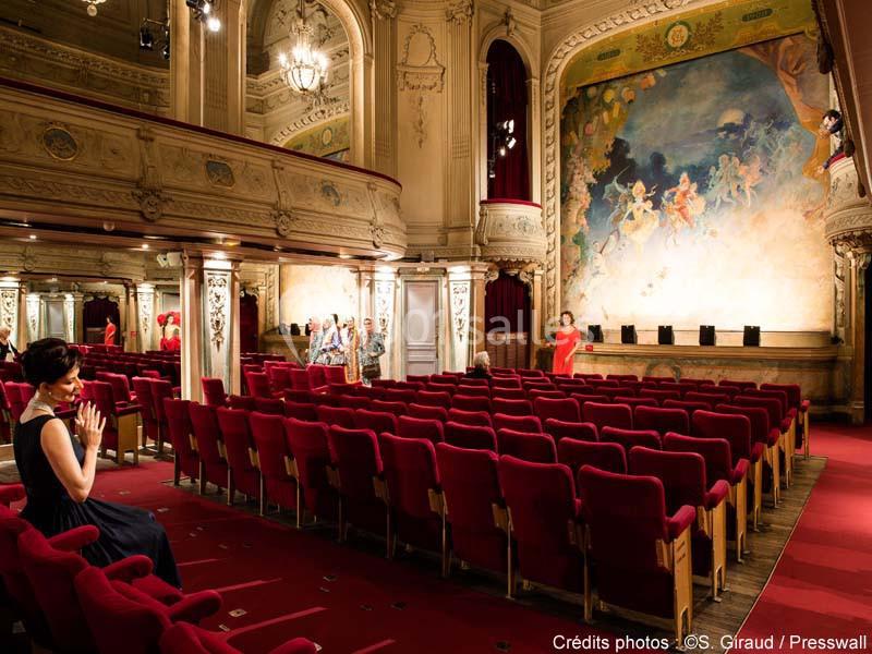 Une femme assise dans une salle de théâtre vide ornée de sièges rouges et d'une fresque murale lumineuse.