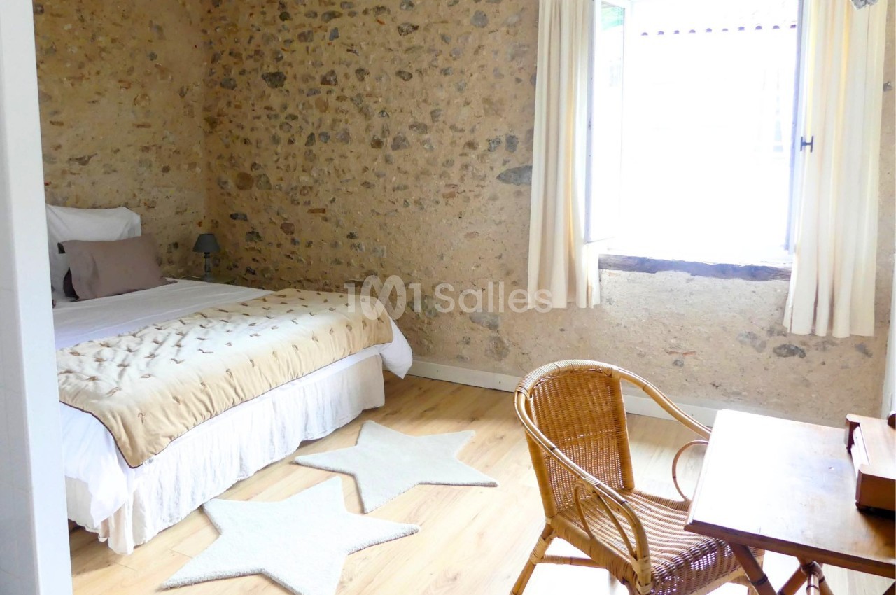 Chambre lumineuse avec murs en pierre, lit double, bureau en bois, chaise en osier et fenêtre avec rideaux clairs.