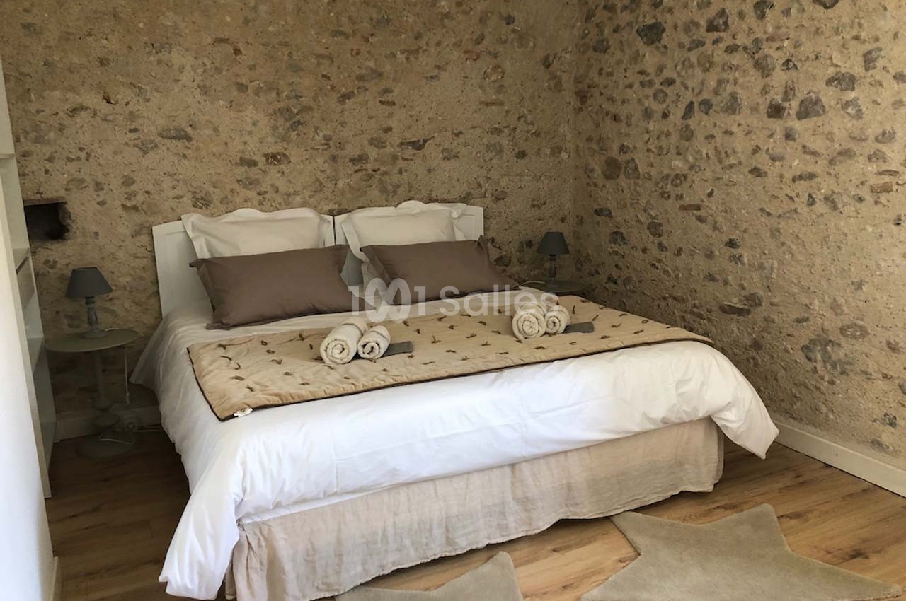 Chambre avec murs en pierre, lit double avec linge beige, coussins assortis et serviettes roulées sur le dessus.