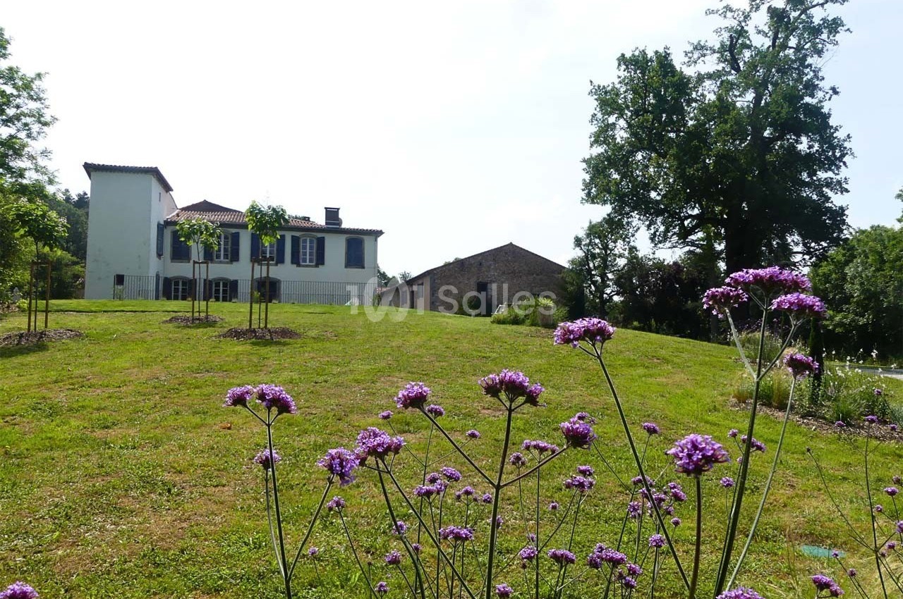 Maison blanche avec volets bleus sur une colline verdoyante, entourée d'arbres et de fleurs violettes au premier plan.