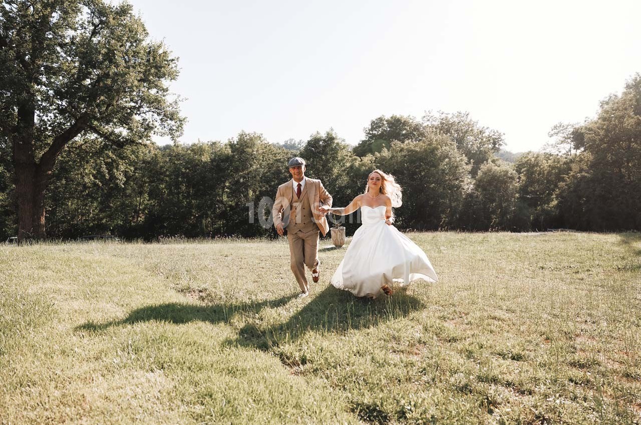 Un couple en tenue de mariage marche main dans la main dans un champ verdoyant sous une lumière naturelle.