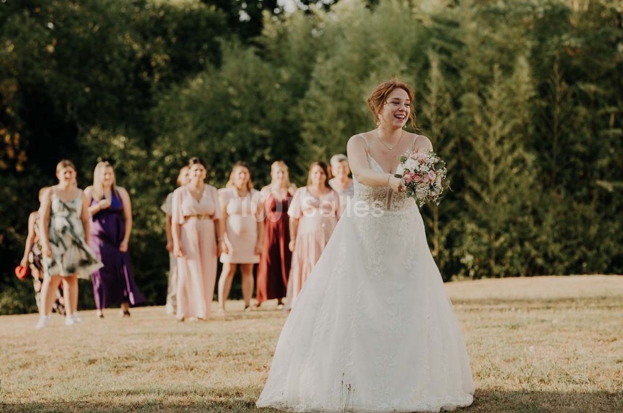 Une mariée en robe blanche lance son bouquet à un groupe de femmes dans un jardin ensoleillé.