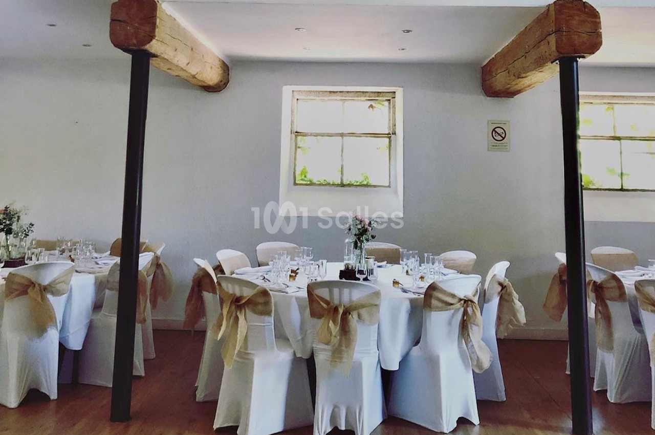 Salle de réception lumineuse avec tables rondes dressées, chaises blanches ornées de nœuds beige, poutres apparentes.