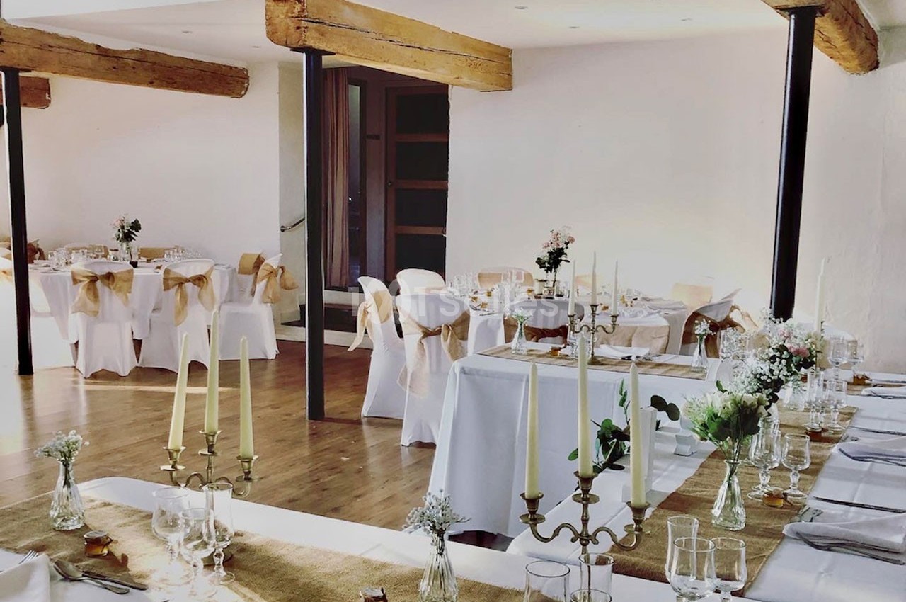 Salle de réception décorée avec des tables dressées, nappes blanches, chandeliers et chaises ornées de rubans beiges.