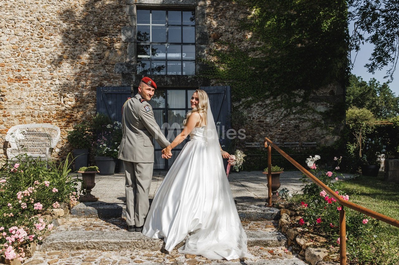 Un couple en tenue de mariage se tient la main devant un bâtiment en pierre entouré de fleurs.