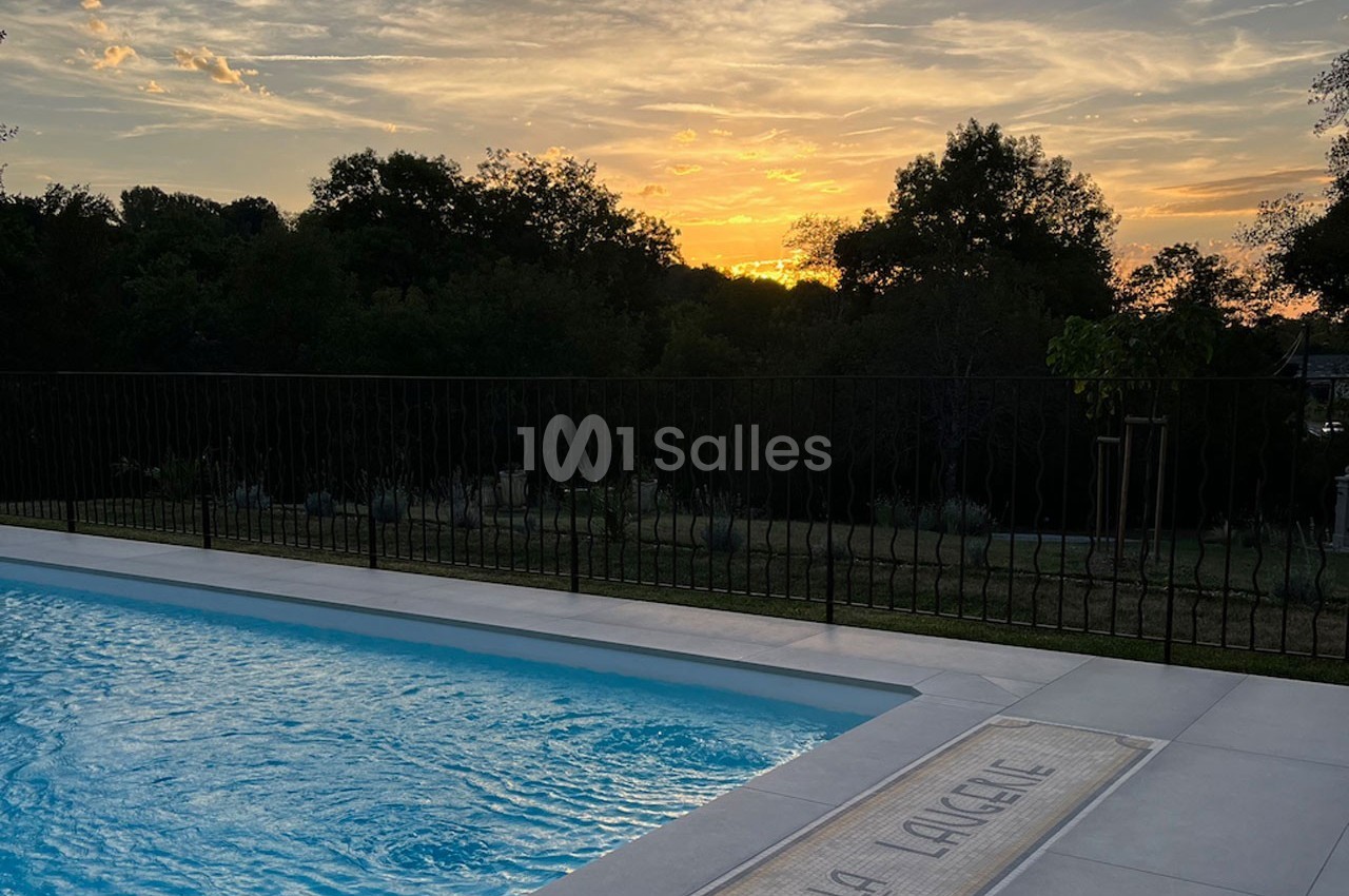 Piscine extérieure avec eau calme au premier plan, entourée de végétation, sous un coucher de soleil.