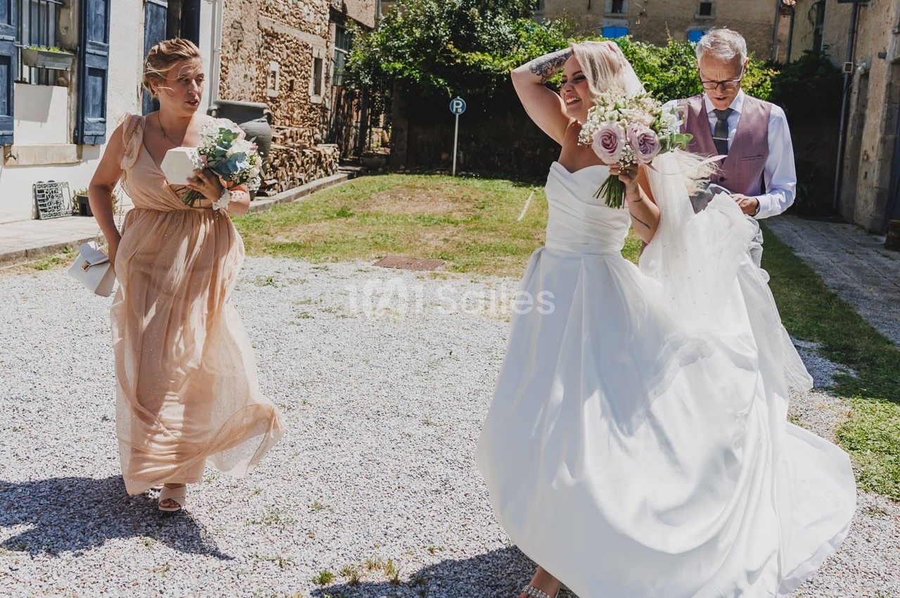 Une mariée en robe blanche marche dehors, accompagnée d'une femme en robe beige et d'un homme en gilet rose.