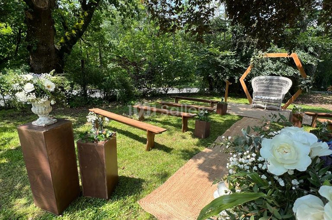 Allée décorée pour une cérémonie en extérieur avec bancs en bois, fleurs blanches et arche géométrique en arrière-plan.