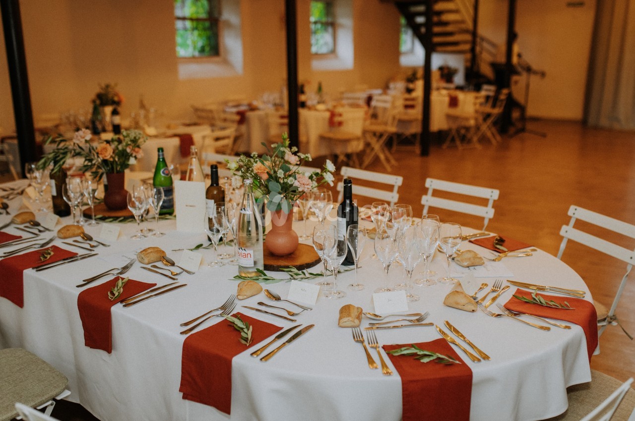 Table ronde décorée pour un repas, avec nappes blanches, chemins de table rouges, vaisselle et arrangements floraux.