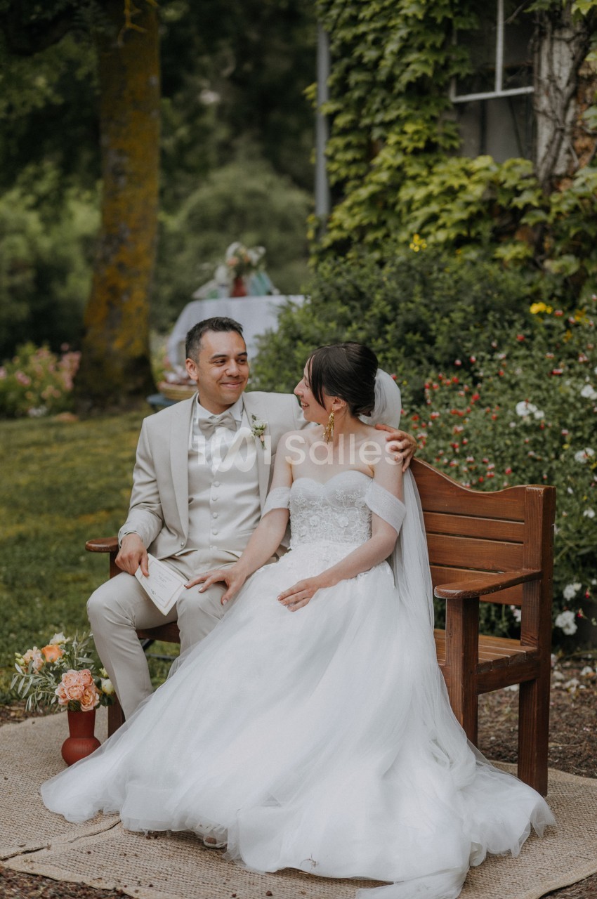 Un couple de mariés assis sur un banc en bois dans un jardin verdoyant, entouré de fleurs.