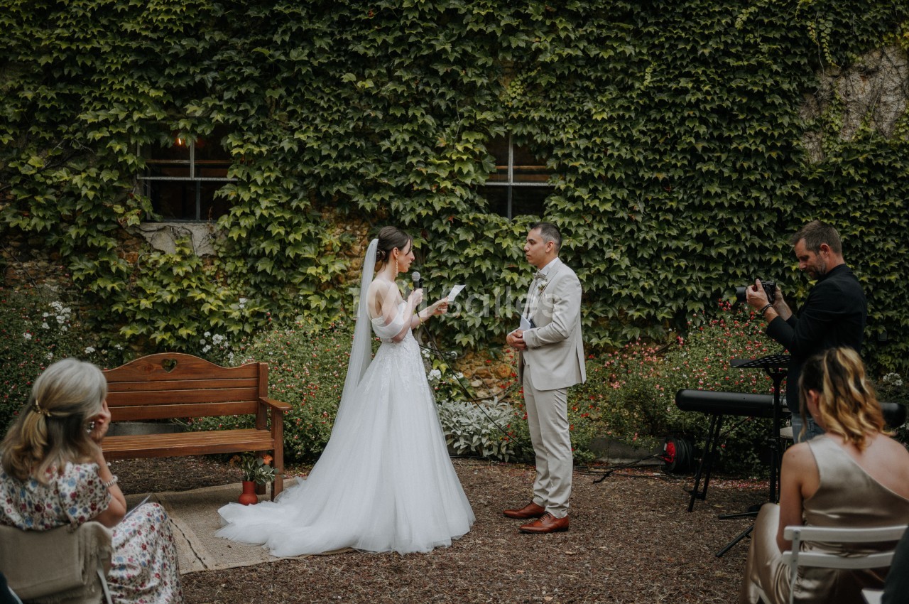 Une mariée lit ses vœux à son époux lors d'une cérémonie en extérieur, entourés de verdure et de quelques invités.