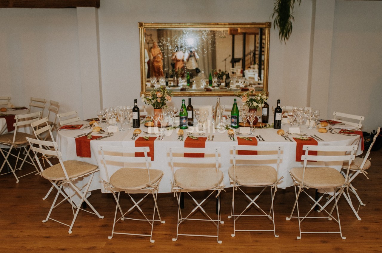 Table dressée pour un repas avec nappes blanches, chaises pliantes, bouteilles, verres et décorations florales.