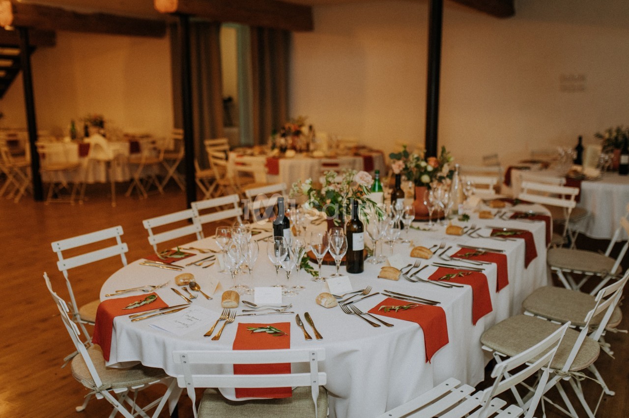 Salle de réception avec tables rondes dressées, nappes blanches, chemins de table rouges et décorations florales.
