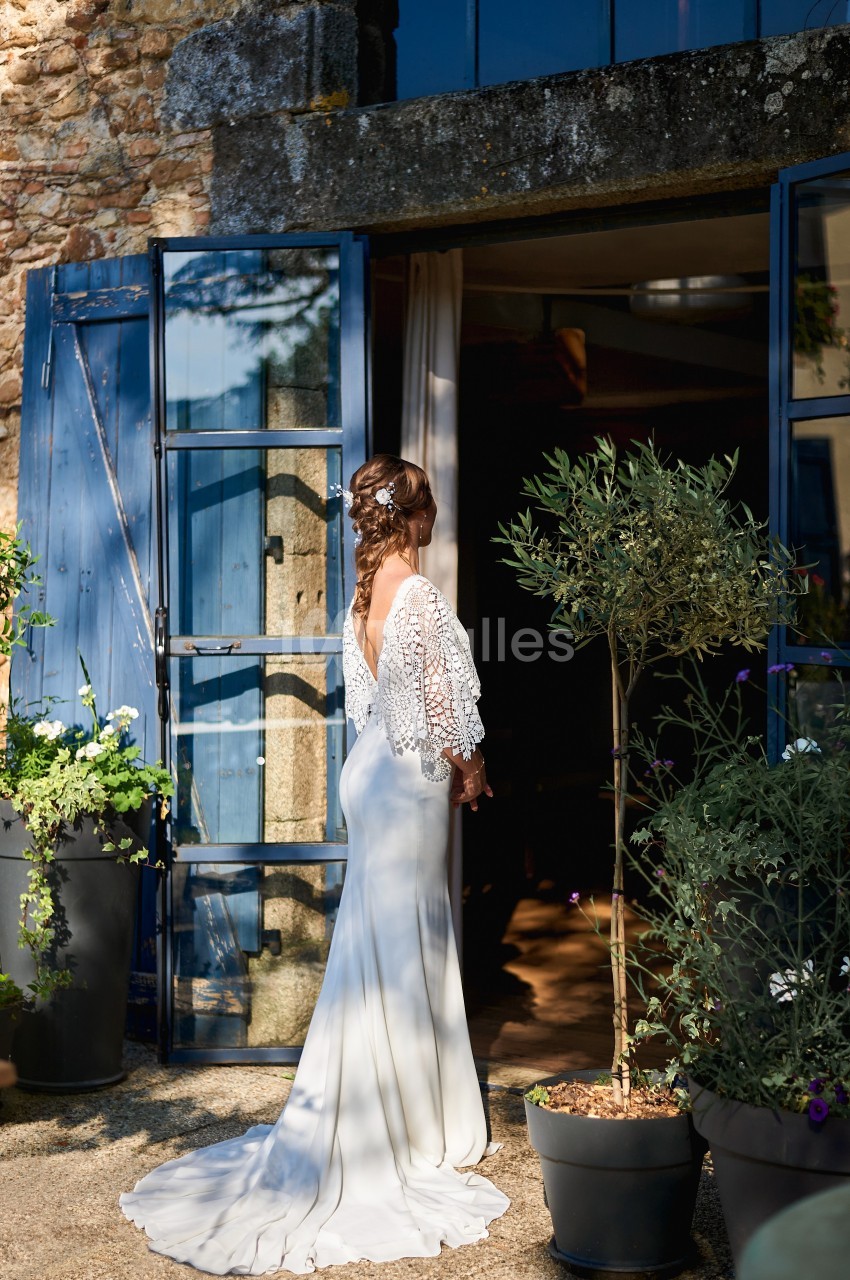 Une femme en robe de mariée blanche se tient devant une porte vitrée ouverte, entourée de plantes en pot.