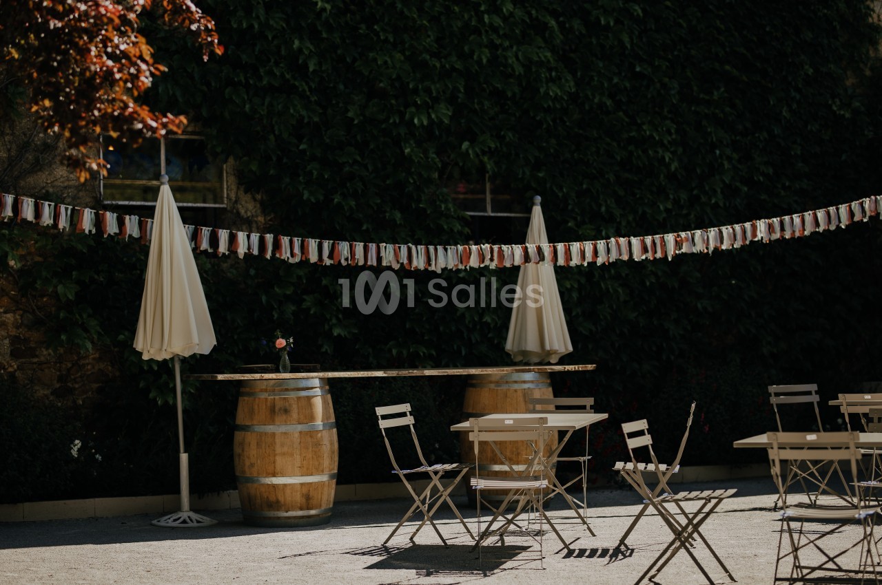 Terrasse extérieure avec tables, chaises pliantes, parasols, tonneaux en bois et guirlande décorative sous un mur végétal.