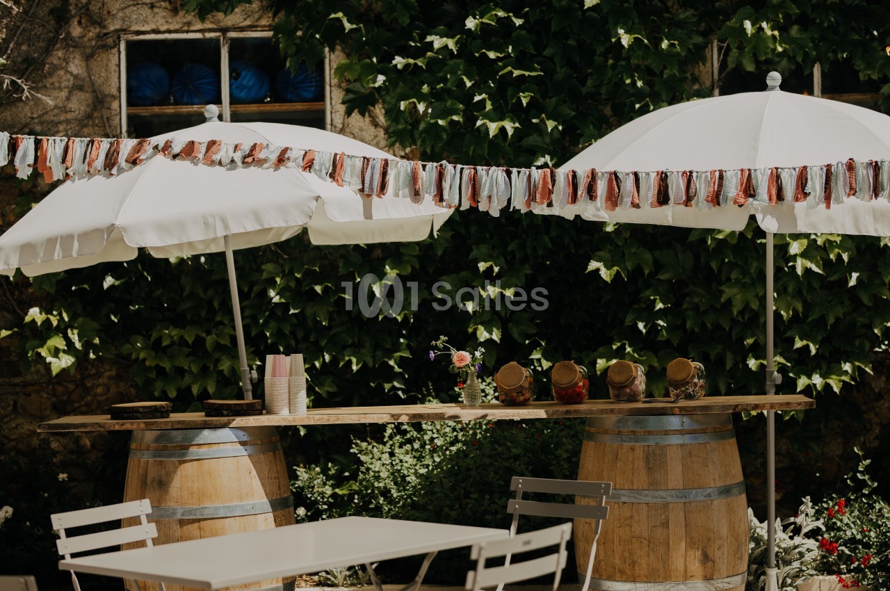 Table en bois posée sur des tonneaux, décorée de pots et guirlandes, sous des parasols blancs dans un jardin verdoyant.