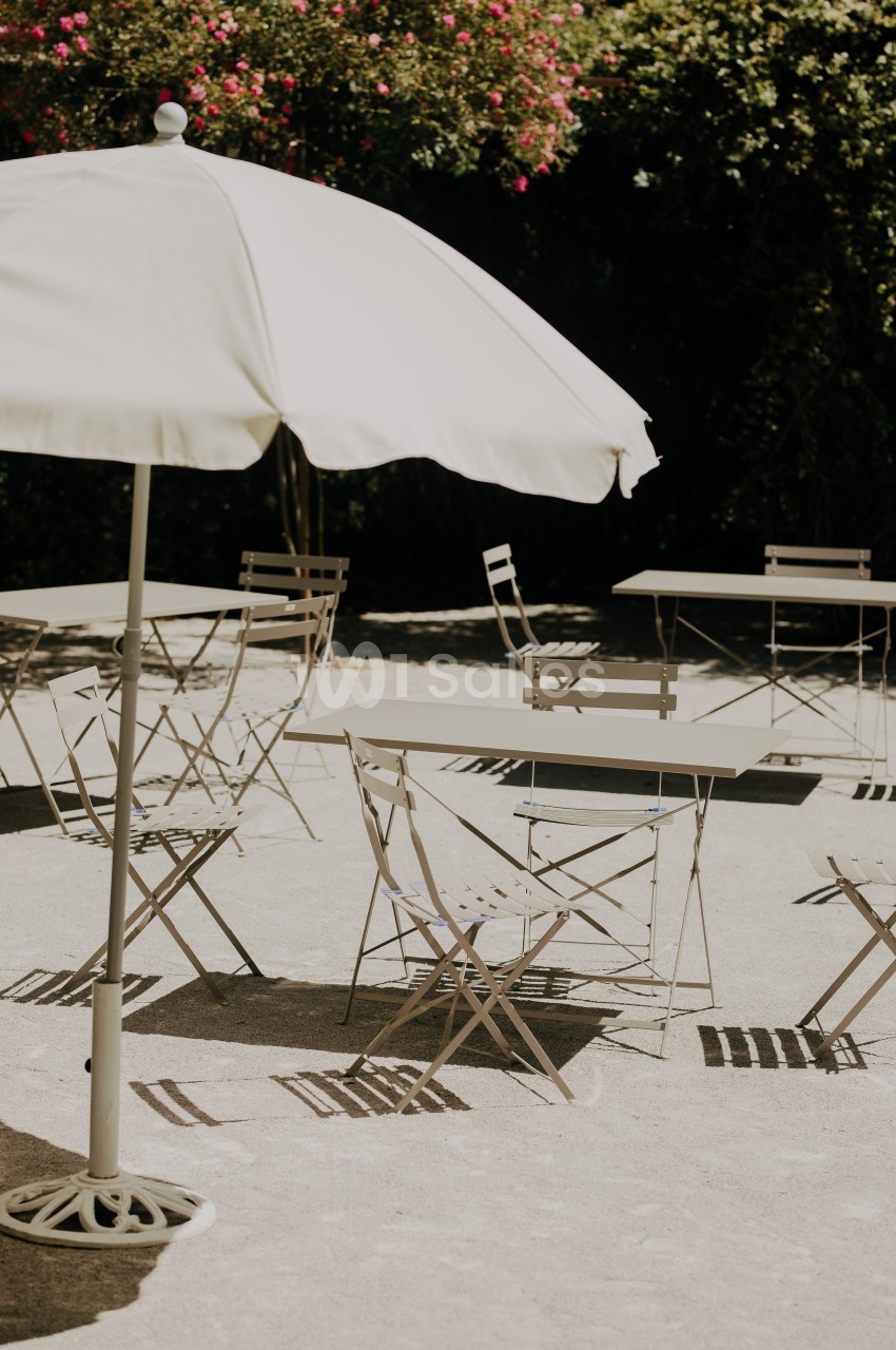 Chaises et tables en métal disposées sous un parasol blanc dans une cour ensoleillée avec des fleurs en arrière-plan.
