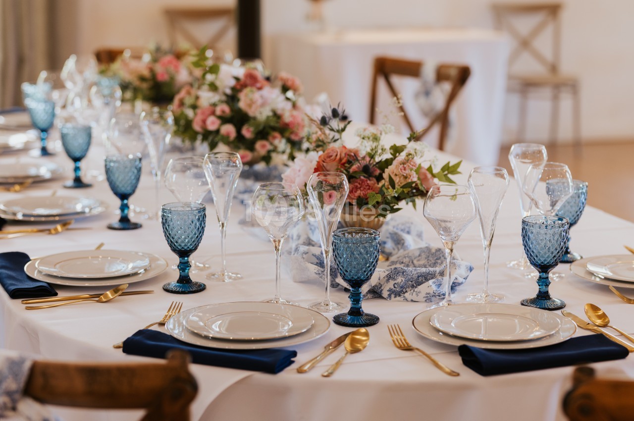 Table élégamment dressée avec vaisselle blanche, verres bleus, couverts dorés et centre de table floral.