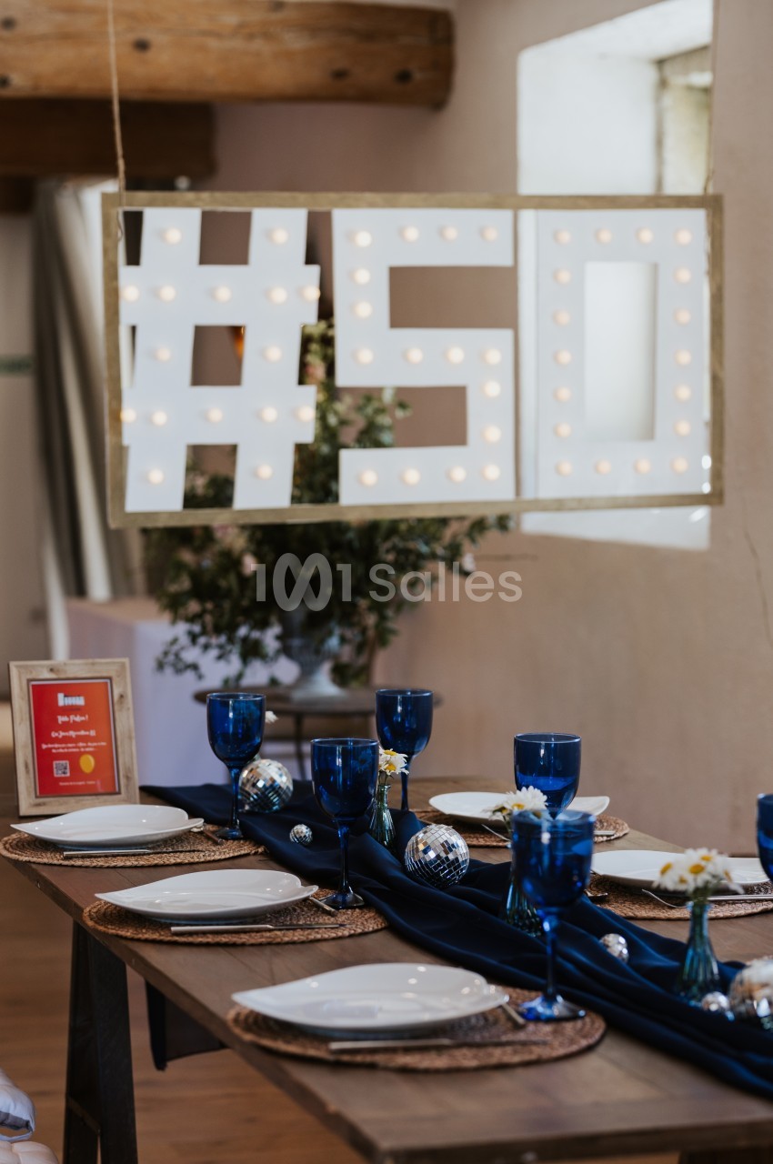 Table décorée avec vaisselle blanche, verres bleus et centre de table sombre, sous un panneau lumineux ’#50’.