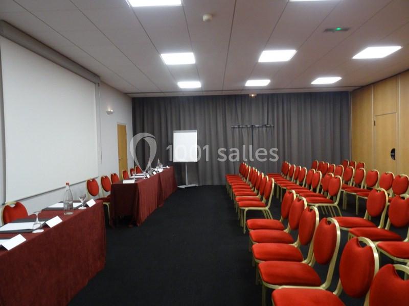 Salle de conférence avec rangées de chaises rouges, tables équipées de micros et écran de projection en arrière-plan.