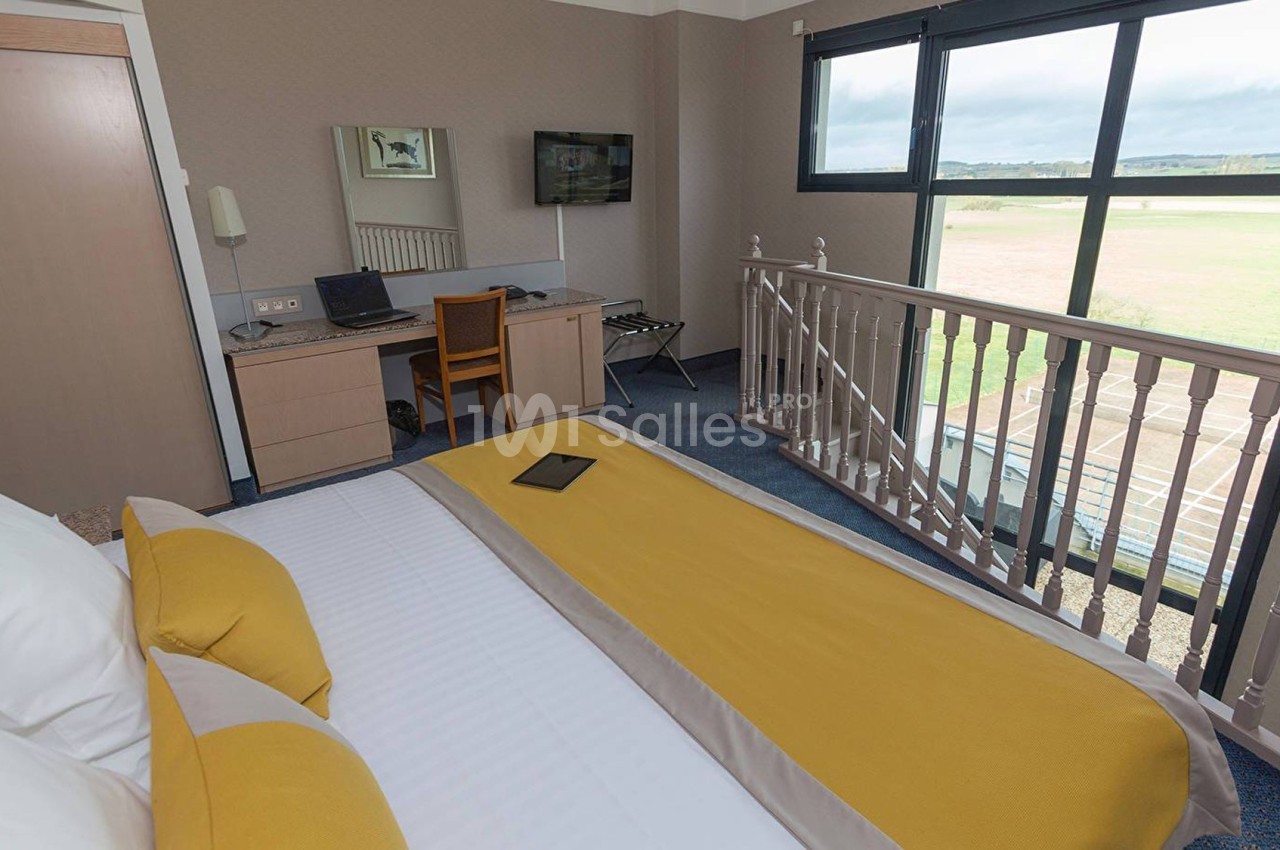 Chambre d'hôtel lumineuse avec lit double, bureau équipé, télévision murale et vue sur un paysage extérieur.