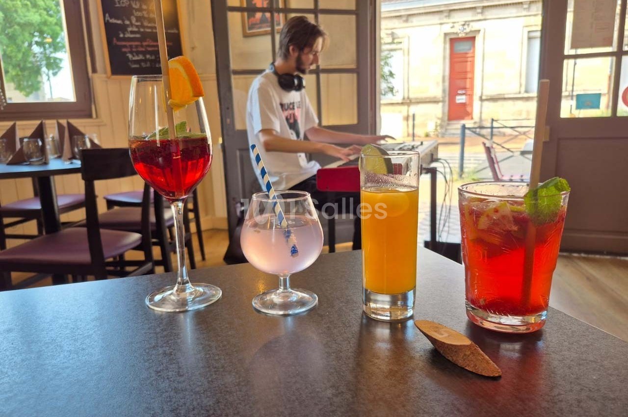 Quatre boissons colorées sur une table dans un café, avec un musicien jouant du clavier en arrière-plan.