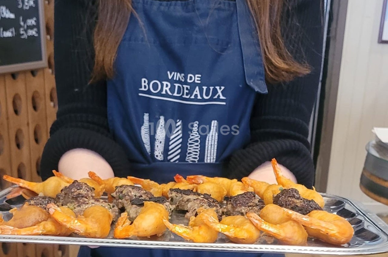 Plateau de bouchées apéritives avec crevettes et viande, tenu par une personne portant un tablier ’Vins de Bordeaux’.