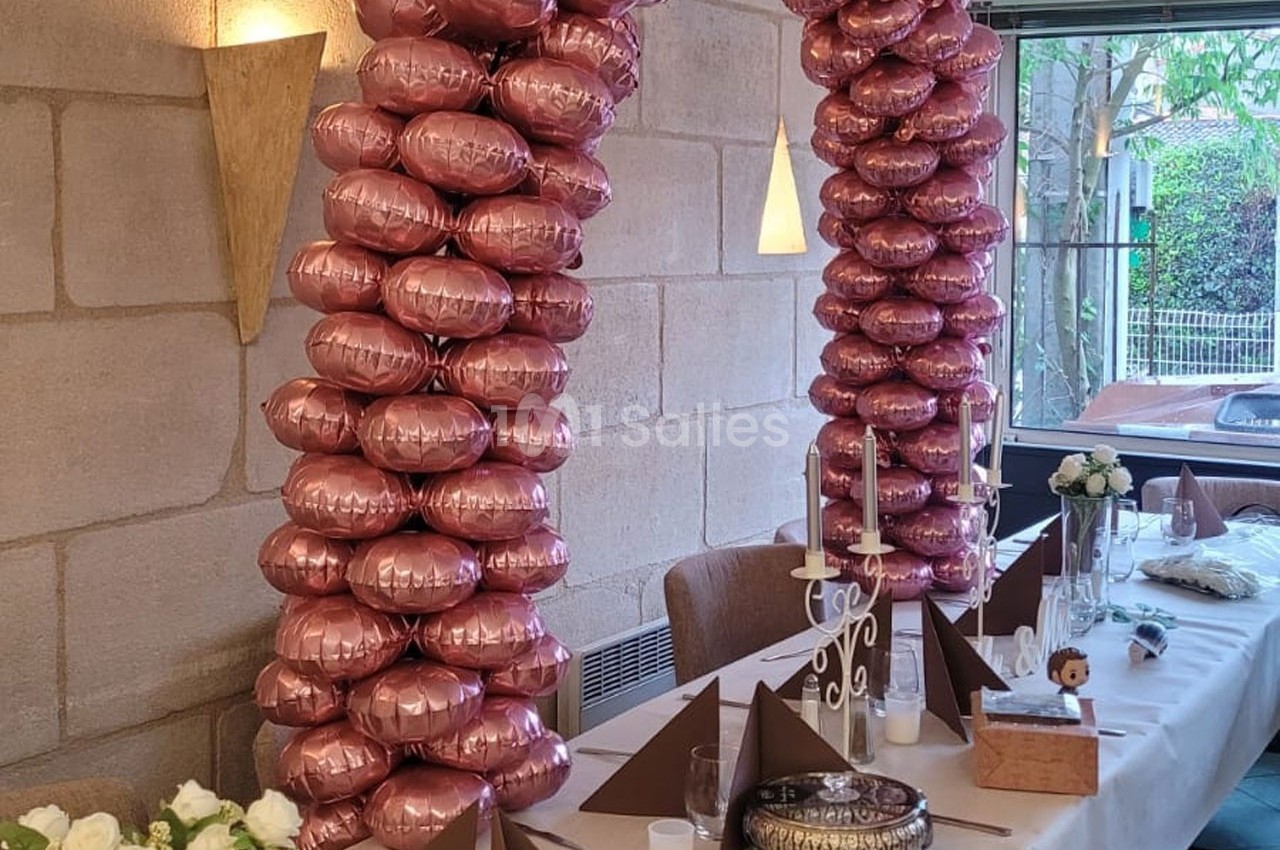Deux colonnes de ballons roses décorent une table de réception avec nappes blanches, bougies et pliages de serviettes.