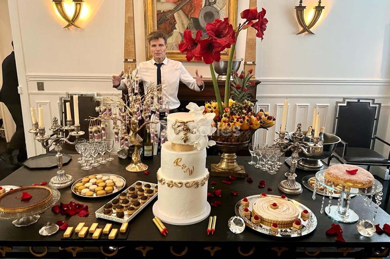 Table décorée avec des desserts variés, un gâteau blanc central, des fleurs rouges et un homme en arrière-plan.