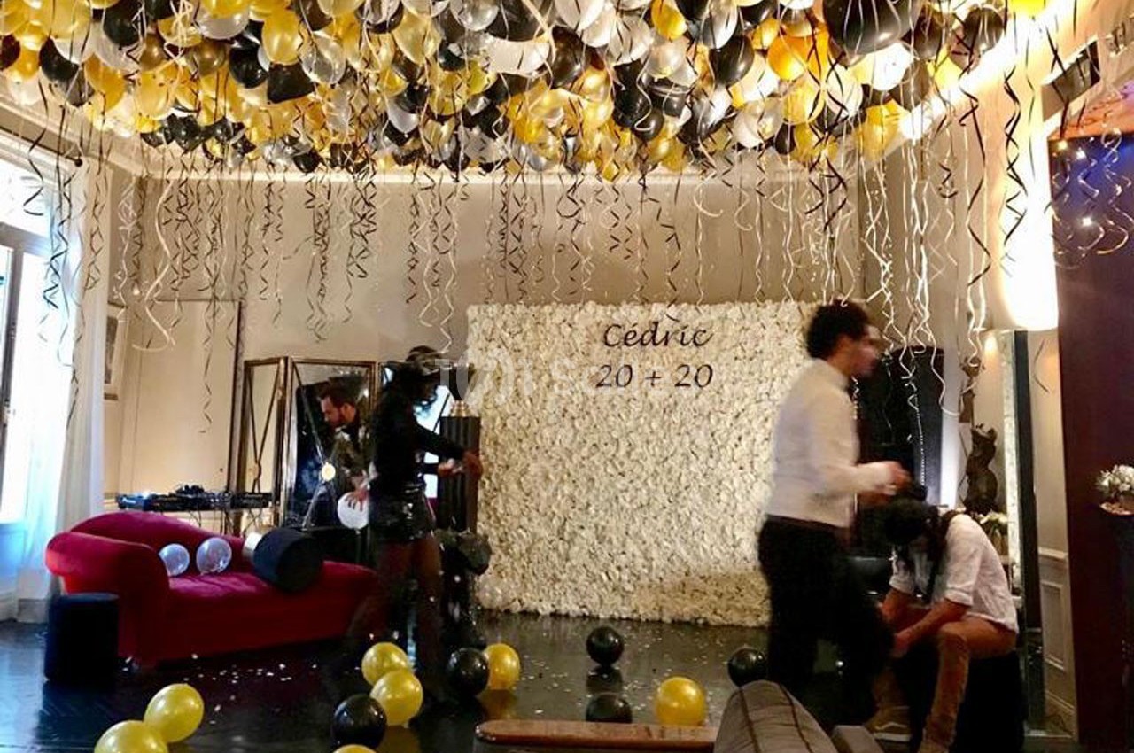 Salle décorée pour une fête avec ballons noirs, dorés et blancs, mur de fleurs blanches et inscription ’Cédric 20 + 20’.