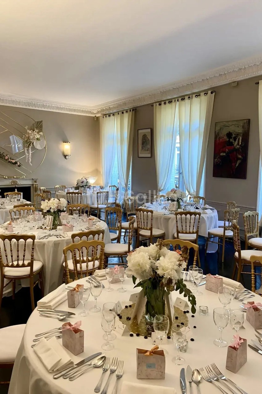 Salle de réception élégamment décorée avec des tables rondes, nappes blanches, chaises dorées et arrangements floraux.