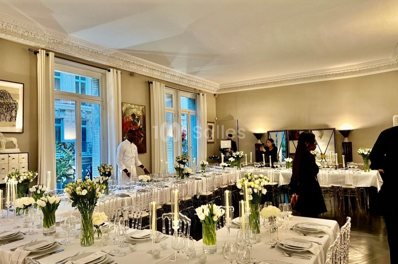 Salle élégante préparée pour un dîner, avec longues tables dressées, chandeliers et bouquets de fleurs blanches.