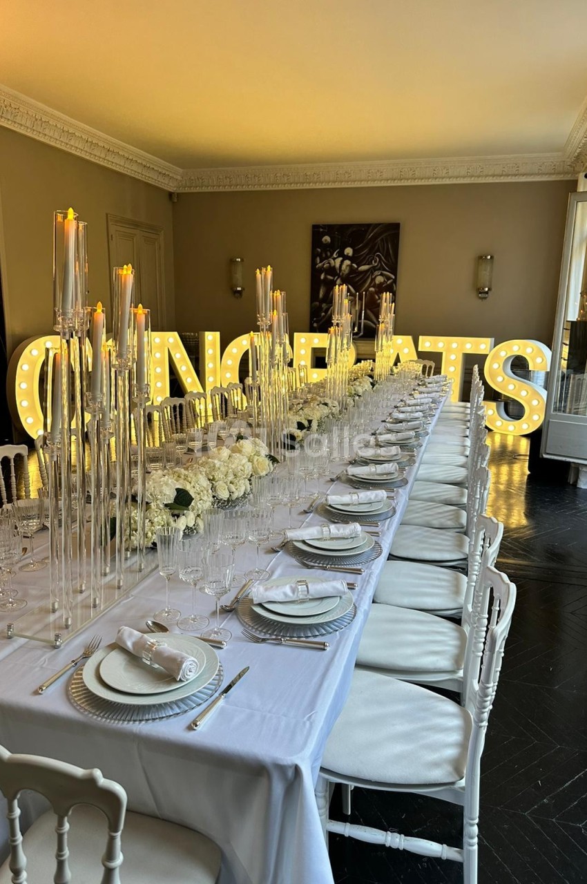 Table élégamment dressée avec chandeliers et fleurs blanches, devant des lettres lumineuses formant ’CONGRATS’.