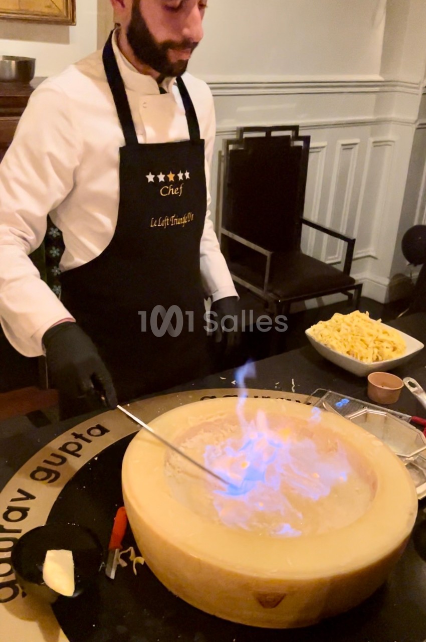 Un chef prépare un plat en flambant du fromage dans une meule, avec des pâtes et des ingrédients à proximité.