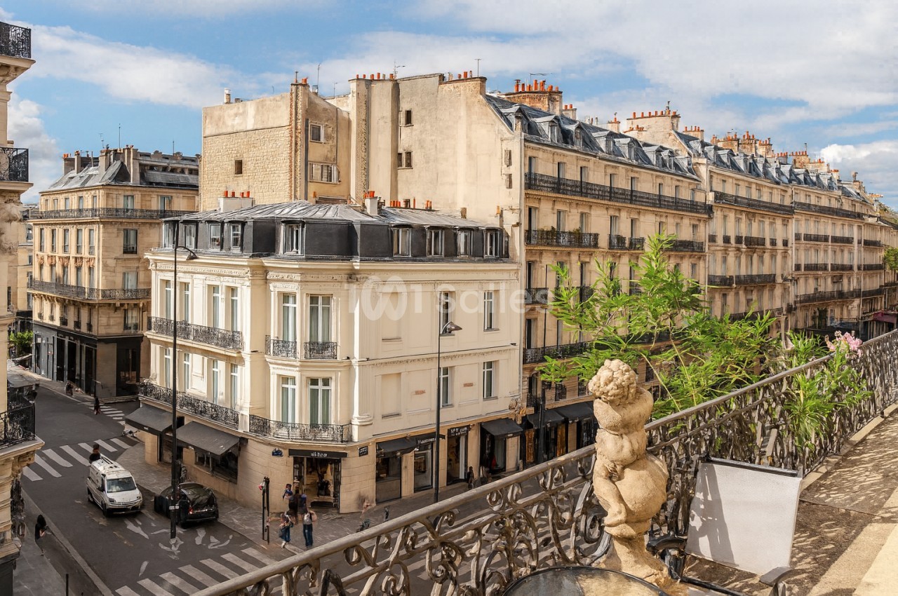 Vue d'une rue parisienne avec des immeubles haussmanniens, un balcon orné et un ciel partiellement nuageux.