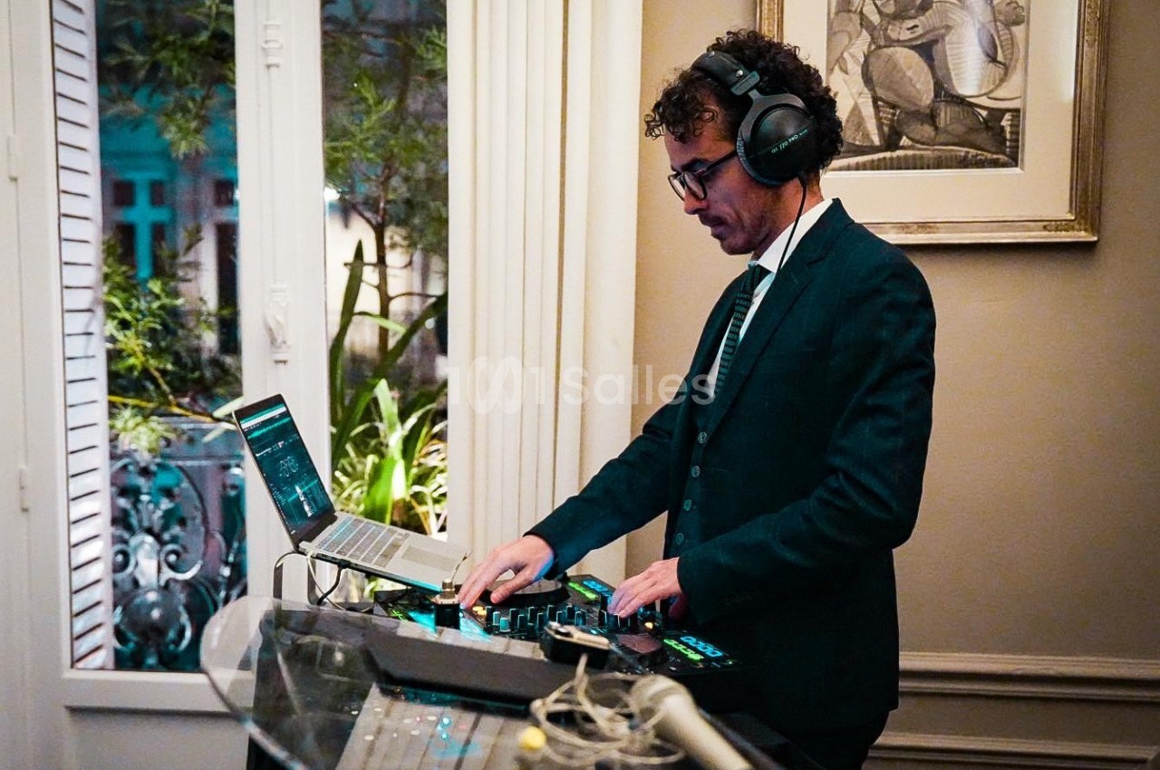 Un DJ en costume mixe de la musique sur une console devant une fenêtre ouverte dans un intérieur élégant.