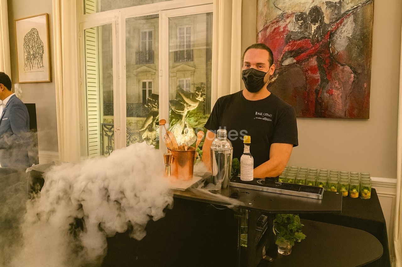 Un barman masqué prépare des cocktails avec de la glace sèche produisant de la fumée, dans un cadre intérieur élégant.