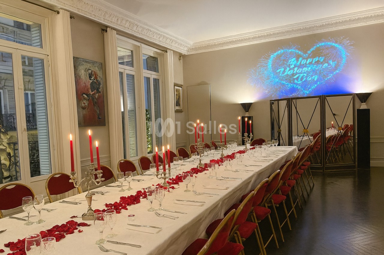 Grande table de banquet décorée de pétales de roses et bougies rouges, dans une salle élégante avec projection murale.
