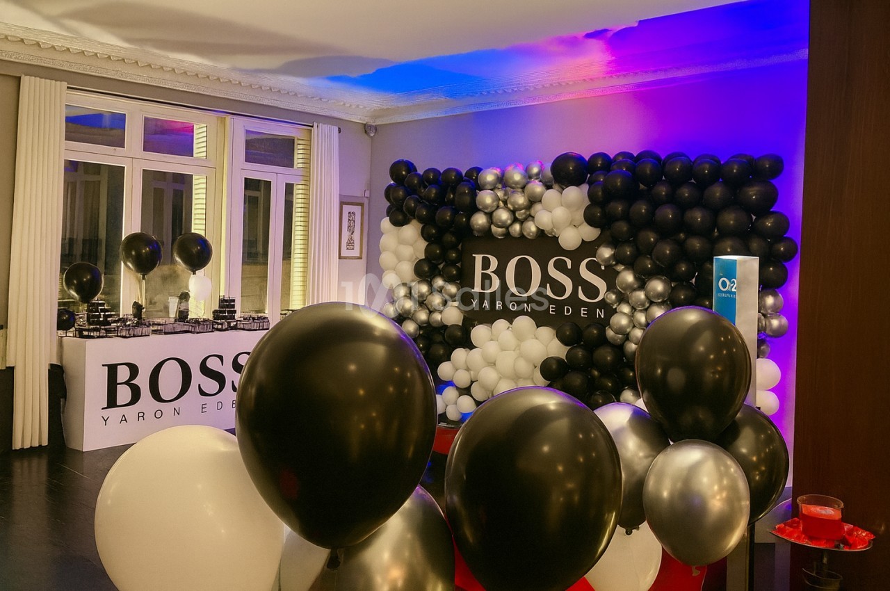 Mur de ballons noirs, blancs et argentés décorant une salle avec une table et un logo ’Boss Yaron Eden’.