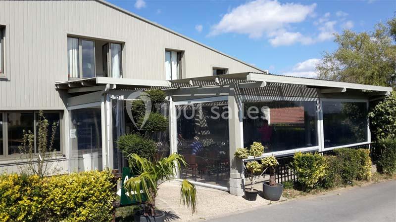 Façade d'un bâtiment avec une terrasse couverte entourée de végétation sous un ciel bleu.