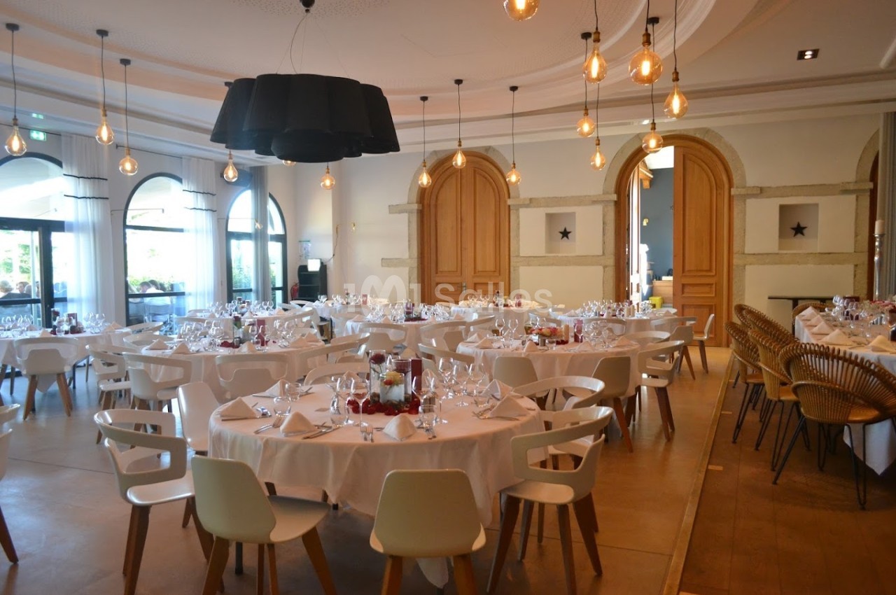 Salle de réception élégante avec tables rondes dressées, chaises blanches et luminaires suspendus.