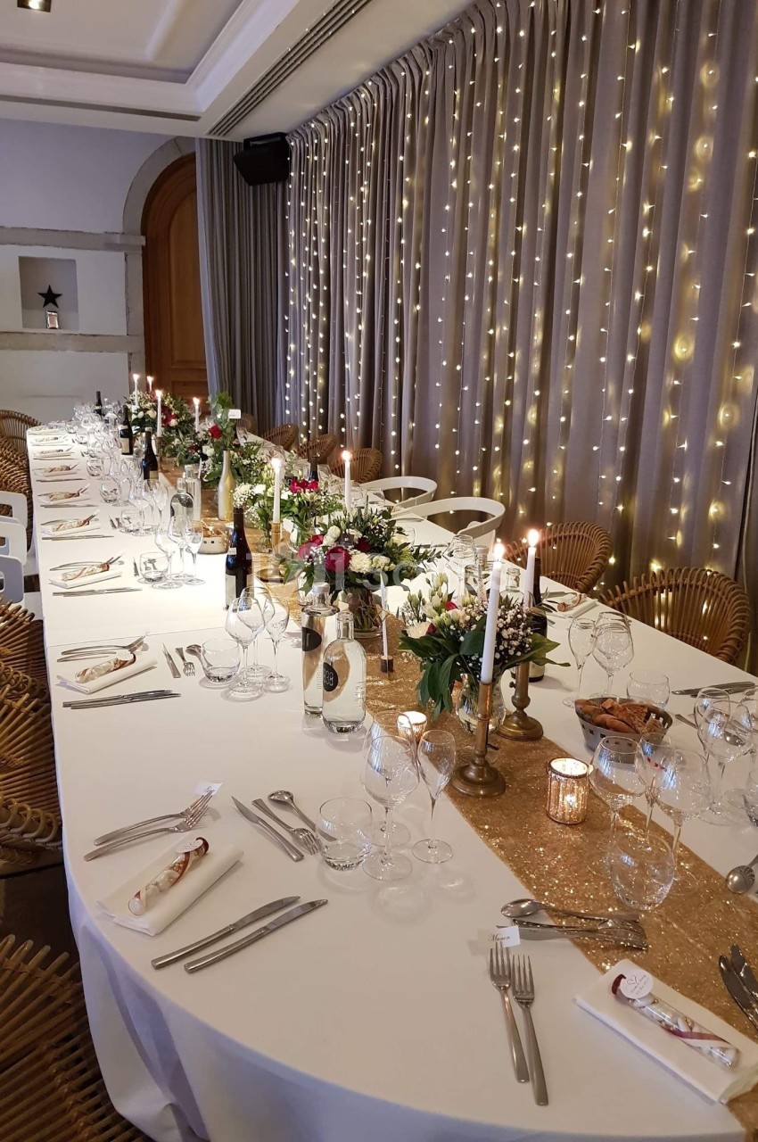 Table élégamment dressée pour un dîner, décorée de bougies, fleurs et guirlandes lumineuses en arrière-plan.