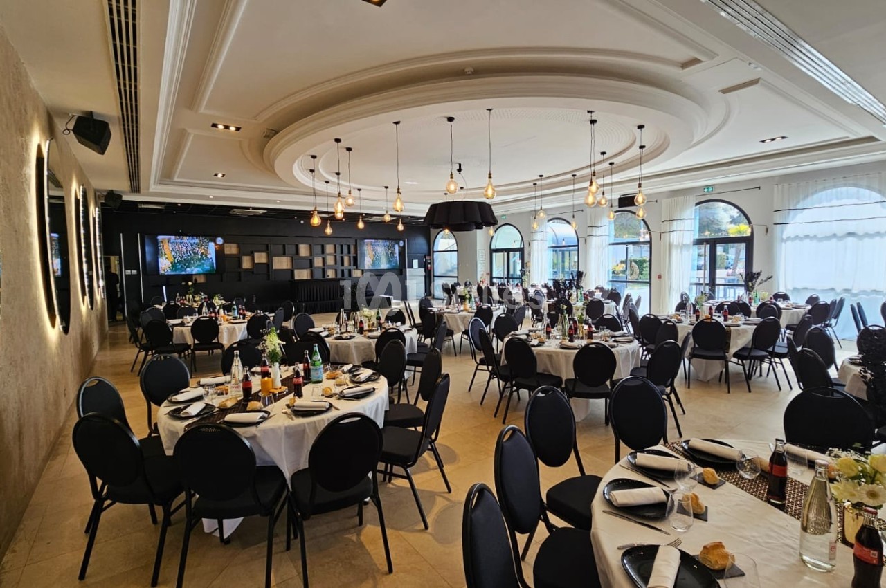 Salle de réception élégante avec tables rondes dressées, chaises noires et grandes baies vitrées laissant entrer la lumière…